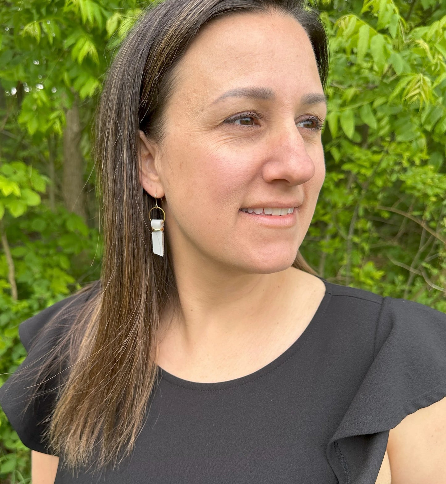 Woman wearing a black top and light gray leather ribbon earrings accented with gold-plated wire and rings, standing outdoors in a green leafy background.