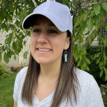 Woman wearing a white cap and blue leather ribbon earrings accented with gold-plated wire and rings, standing outdoors with greenery in the background