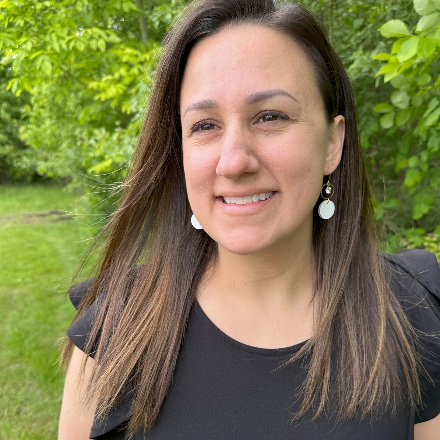 Woman with long brown hair wearing a black shirt and white leather statement earrings featuring gold-plated rings and crystal details, smiling outdoors with greenery in the background.