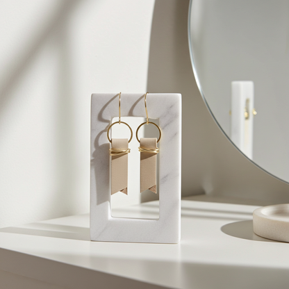 Beige leather earrings accented with gold-plated rings and wire displayed in a white marble box on a light surface with a mirror in the background.