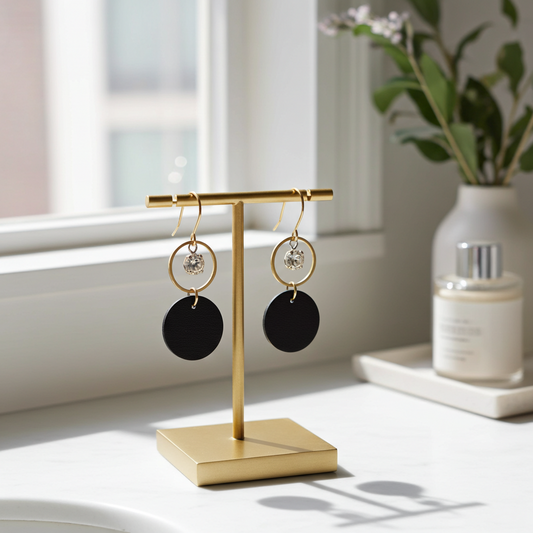 A pair of black leather statement earrings featuring gold-plated rings and crystal details on a gold earring stand sitting on a white surface with a window in the background.