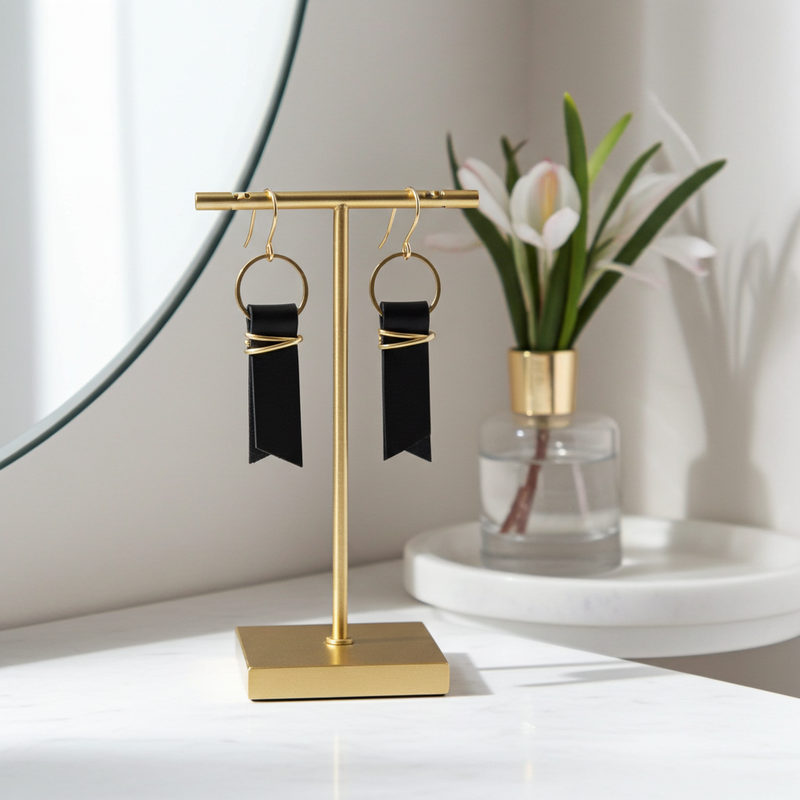 Gold earring stand with black leather ribbon earrings accented with gold-plated wire and rings on a white surface with a mirror and plant in the background.