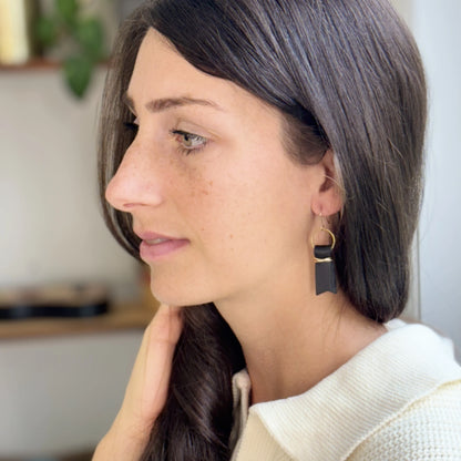 A woman indoors wearing a white sweater and black leather ribbon earrings accented with gold-plated wire and rings. 