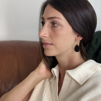 Woman wearing black leather teardrop earrings with gold-plated hooks sitting on a couch.