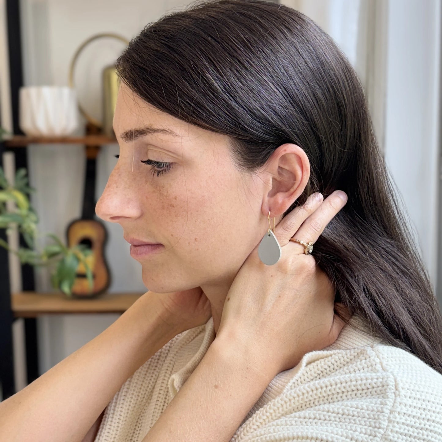 Woman wearing light gray teardrop earrings with gold-plated hooks in a home setting.