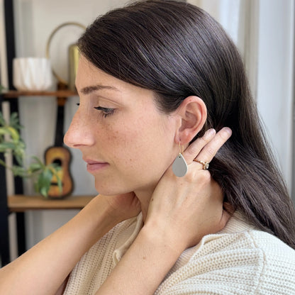 Woman wearing light gray teardrop earrings with gold-plated hooks in a home setting.