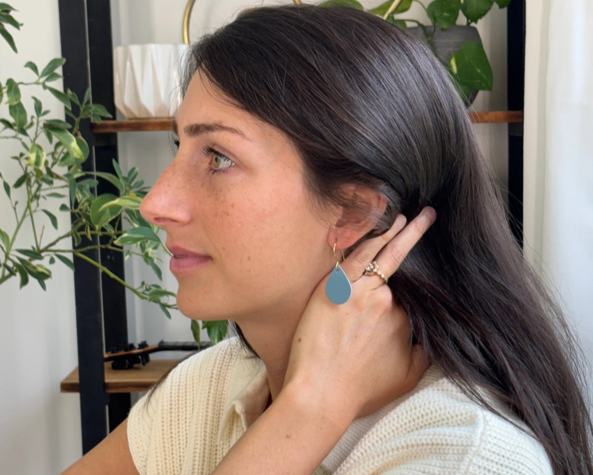 Woman wearing blue leather teardrop earrings with gold-plated hooks, sitting in a home setting with plants and decor.