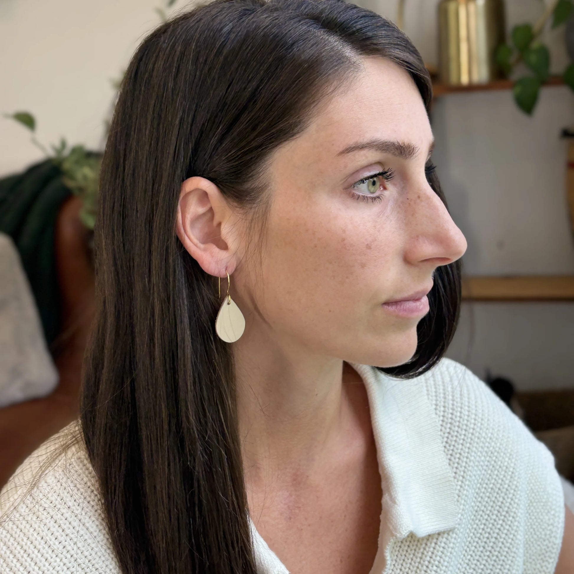 Woman wearing beige teardrop earrings with gold-plated hooks, sitting in a casual indoor setting
