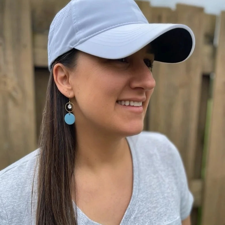Woman wearing a white cap, blue leather statement earrings featuring gold-plated rings and crystal details, and a gray shirt standing outdoors with a wooden fence background.