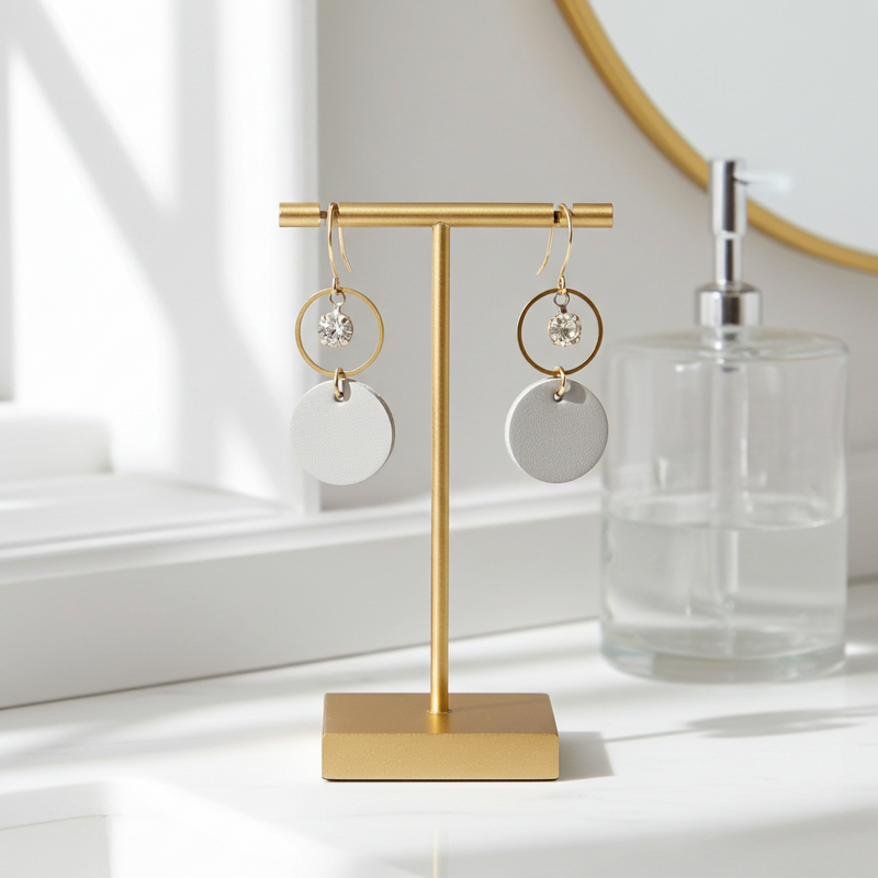 Gold earring stand with light gray leather statement earrings featuring gold-plated rings and crystal details on a white surface.
