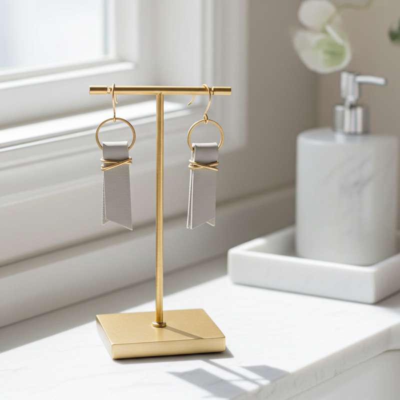 A pair of light gray leather ribbon earrings accented with gold-plated wire and rings on a gold earring stand sitting on a white surface.