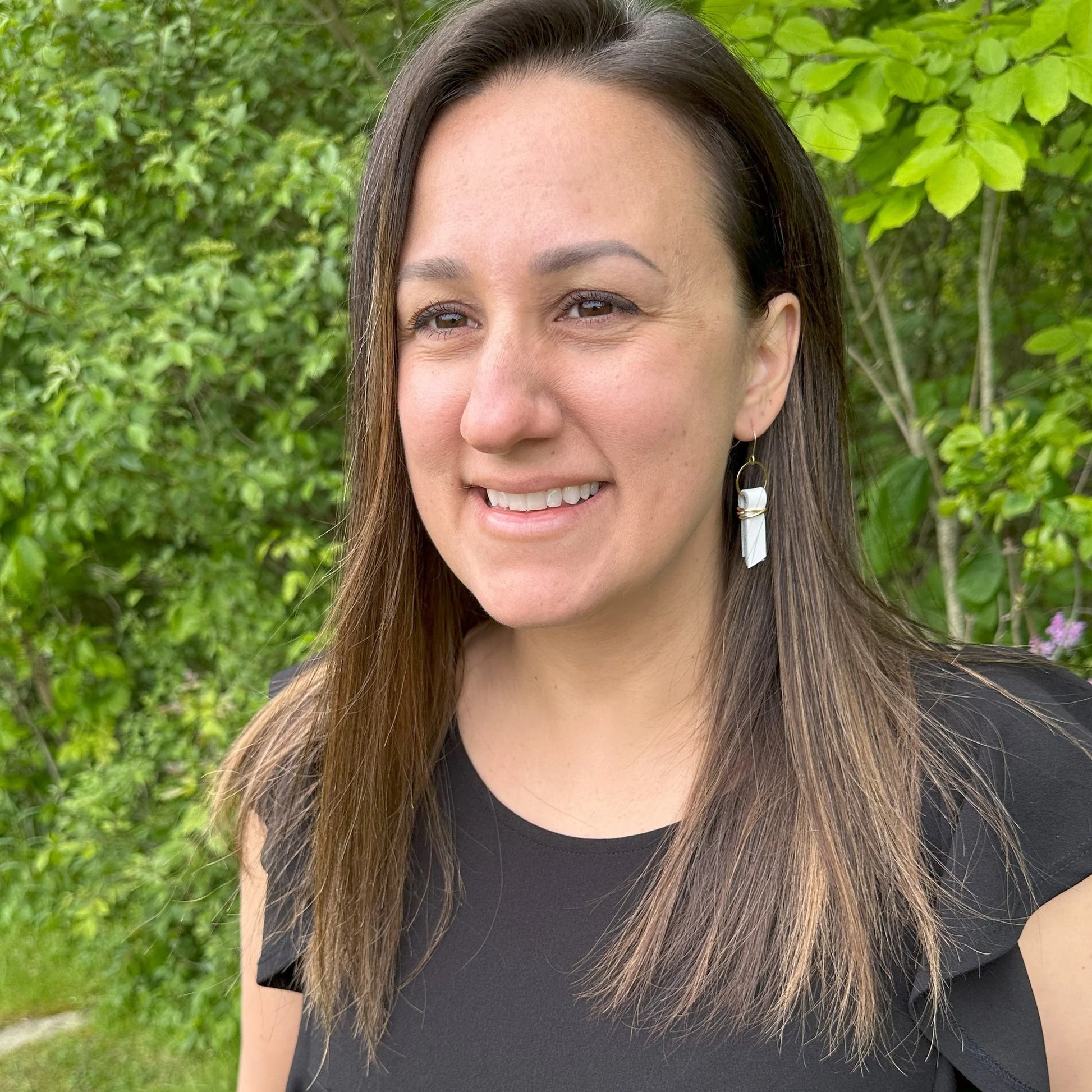 Woman with long brown hair wearing a black top and white leather ribbon earrings accented with gold-plated wire and rings, standing outdoors with greenery in the background