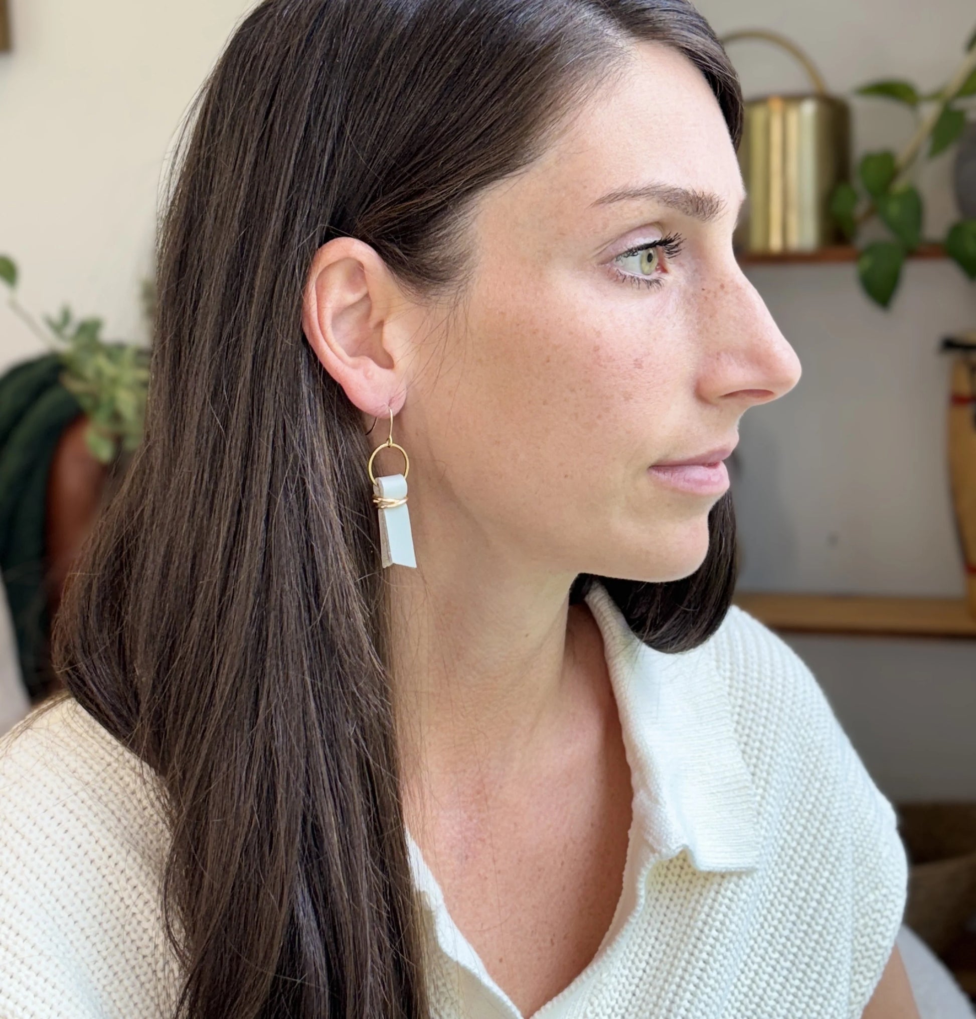 A woman indoors wearing a white sweater and seafoam green leather ribbon earrings accented with gold-plated wire and rings. 