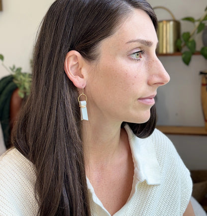 A woman indoors wearing a white sweater and seafoam green leather ribbon earrings accented with gold-plated wire and rings. 