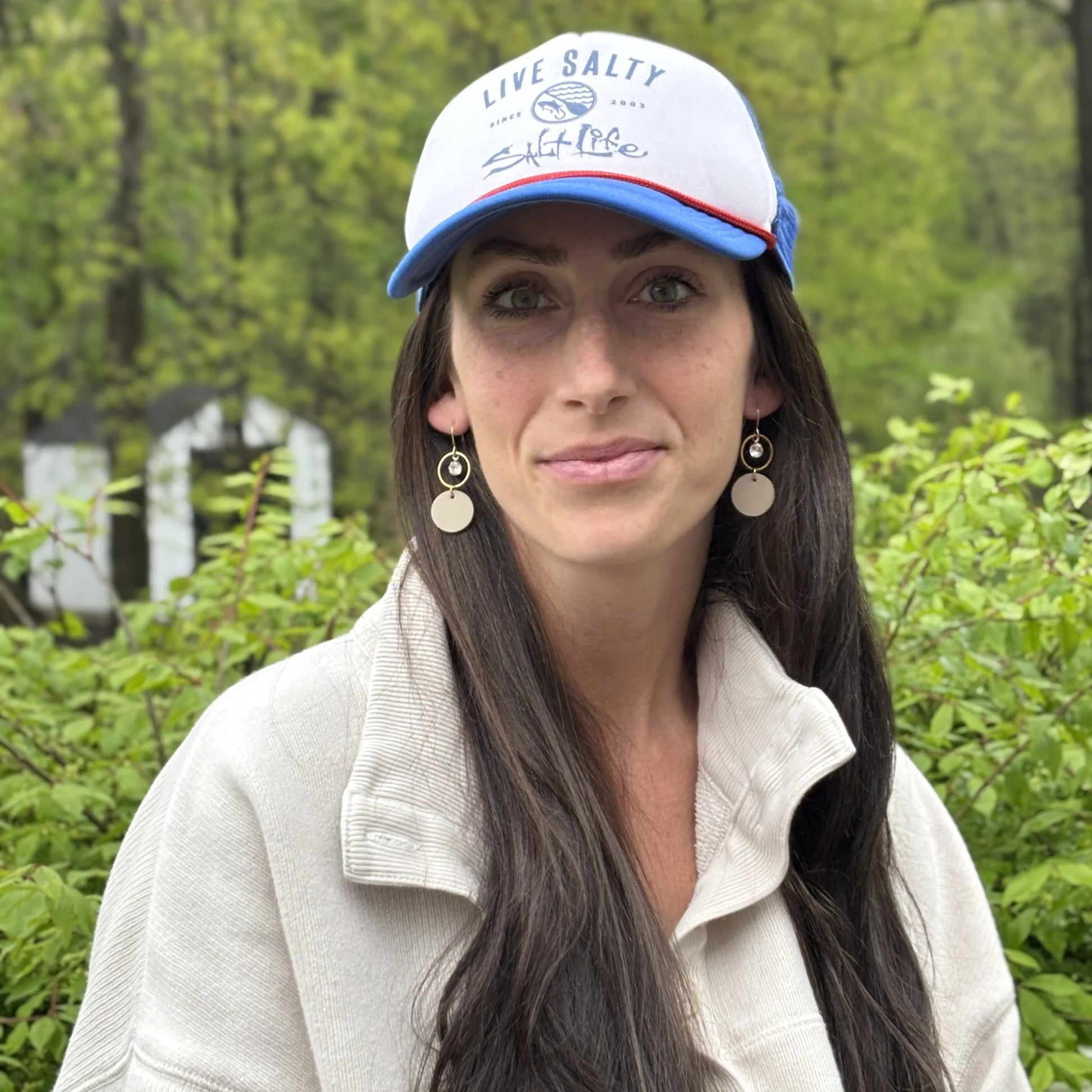 Woman wearing a cap with 'Live Salty' text and beige leather statement earrings featuring gold-plated rings and crystal details, seated outdoors.