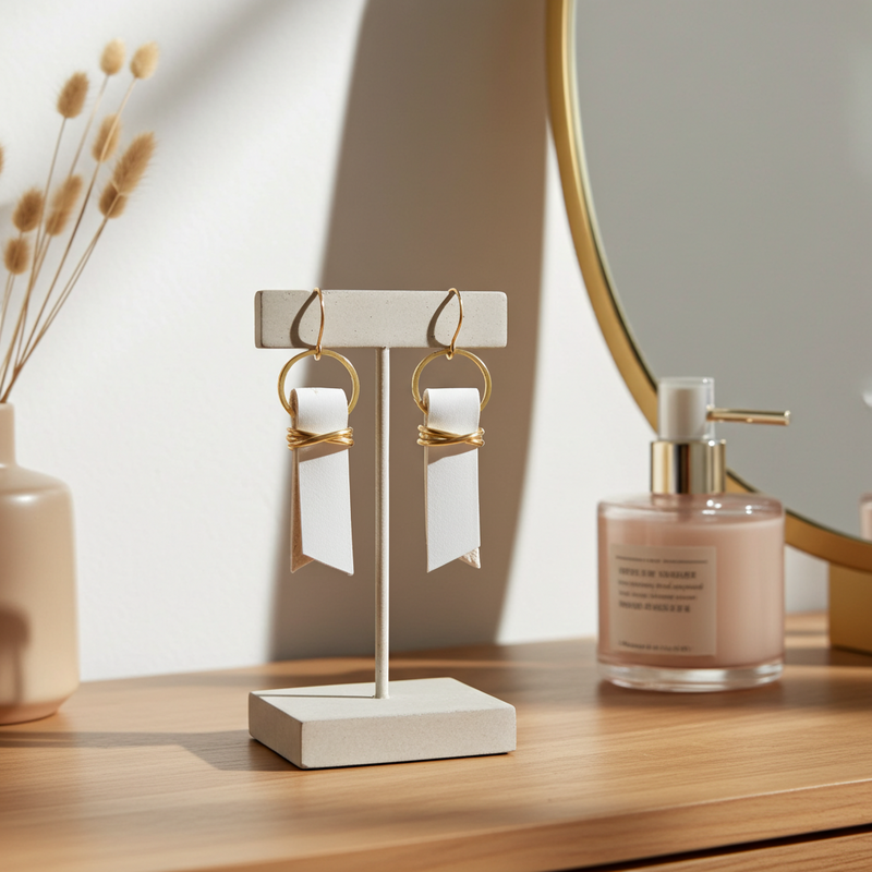 Off-white leather ribbon earrings accented with gold-plated wire and rings on a stand with a bottle of perfume and a vase in the background.