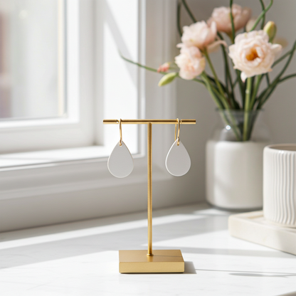 Off-white leather teardrop earrings with gold-plated hooks on a gold stand with a vase of flowers in the background.