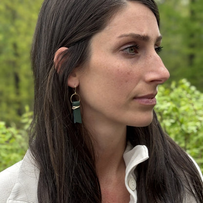 Woman wearing cream sweater and green leather ribbon earrings accented with gold-plated wire and rings, standing in a blurred outdoor setting with trees. 