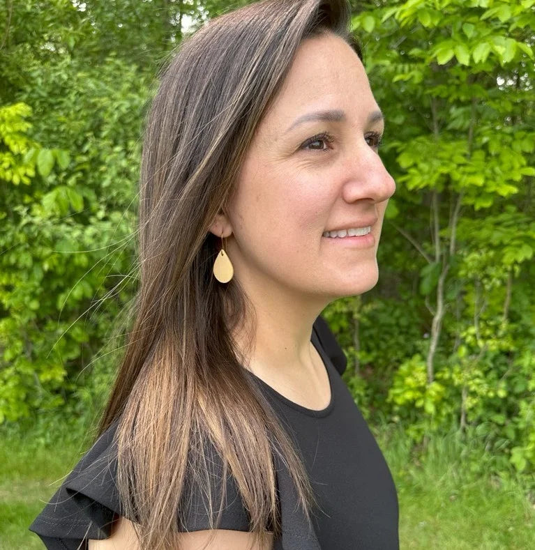 Woman with long brown hair wearing yellow leather teardrop earrings with gold-plated hooks and a black top outdoors with greenery in the background.