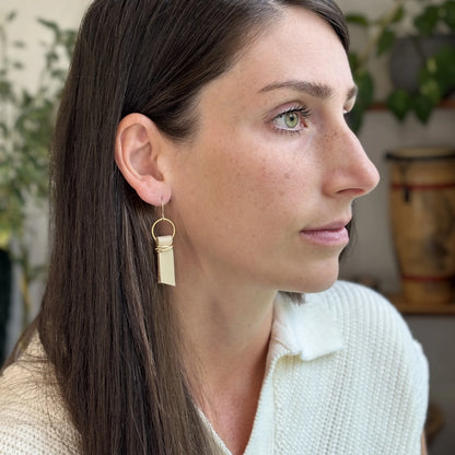 A woman indoors wearing a white sweater and beige leather ribbon earrings accented with gold-plated wire and rings. 
