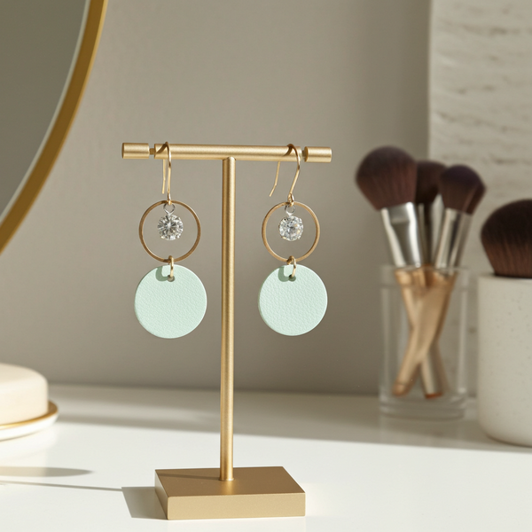 Gold earring stand with seafoam green leather earrings accented with crystals inside gold-plated rings on a neutral background
