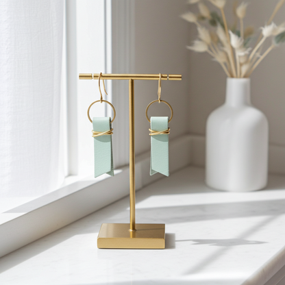 Gold earring stand with seafoam green leather earrings in a bright room with a vase in the background.