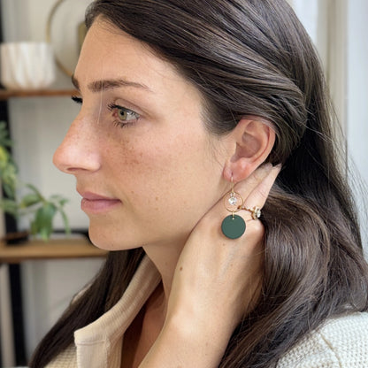 Woman wearing green leather statement earrings featuring gold-plated rings and crystal details, sitting in a blurred indoor background