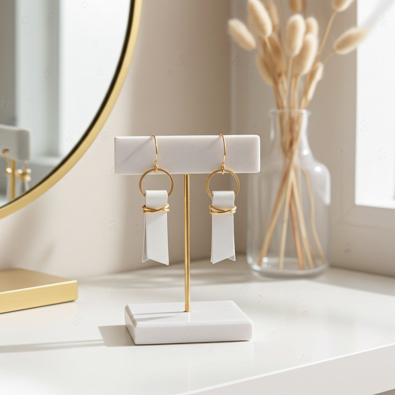 White leather ribbon earrings accented with gold-plated wire and rings on a marble stand with a vase of dried plants in the background.