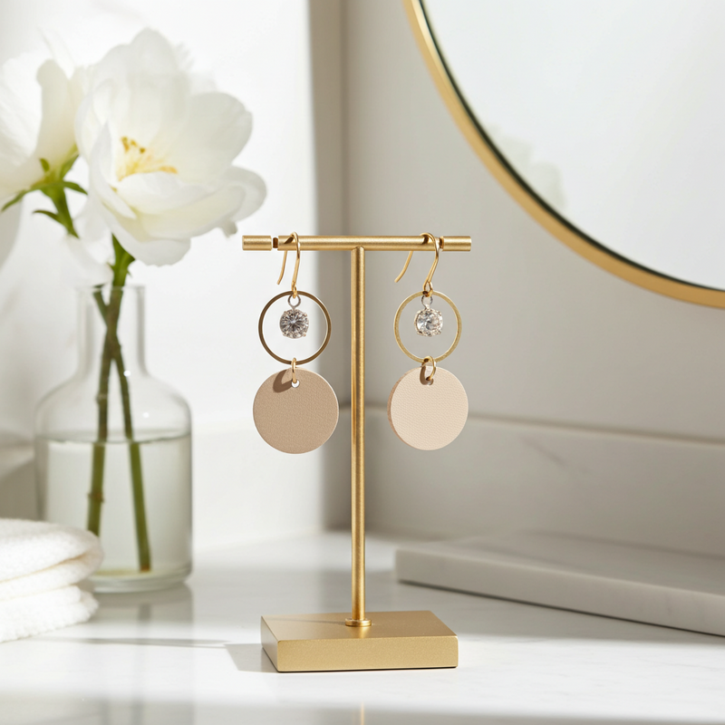 Gold earring stand with beige leather statement earrings featuring gold-plated rings and crystal details on a white surface with flowers in the background